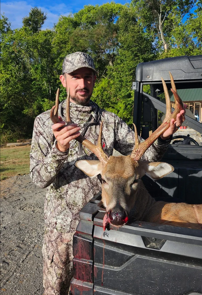 Hunter in camouflage standing beside a buck in a pickup truck bed.