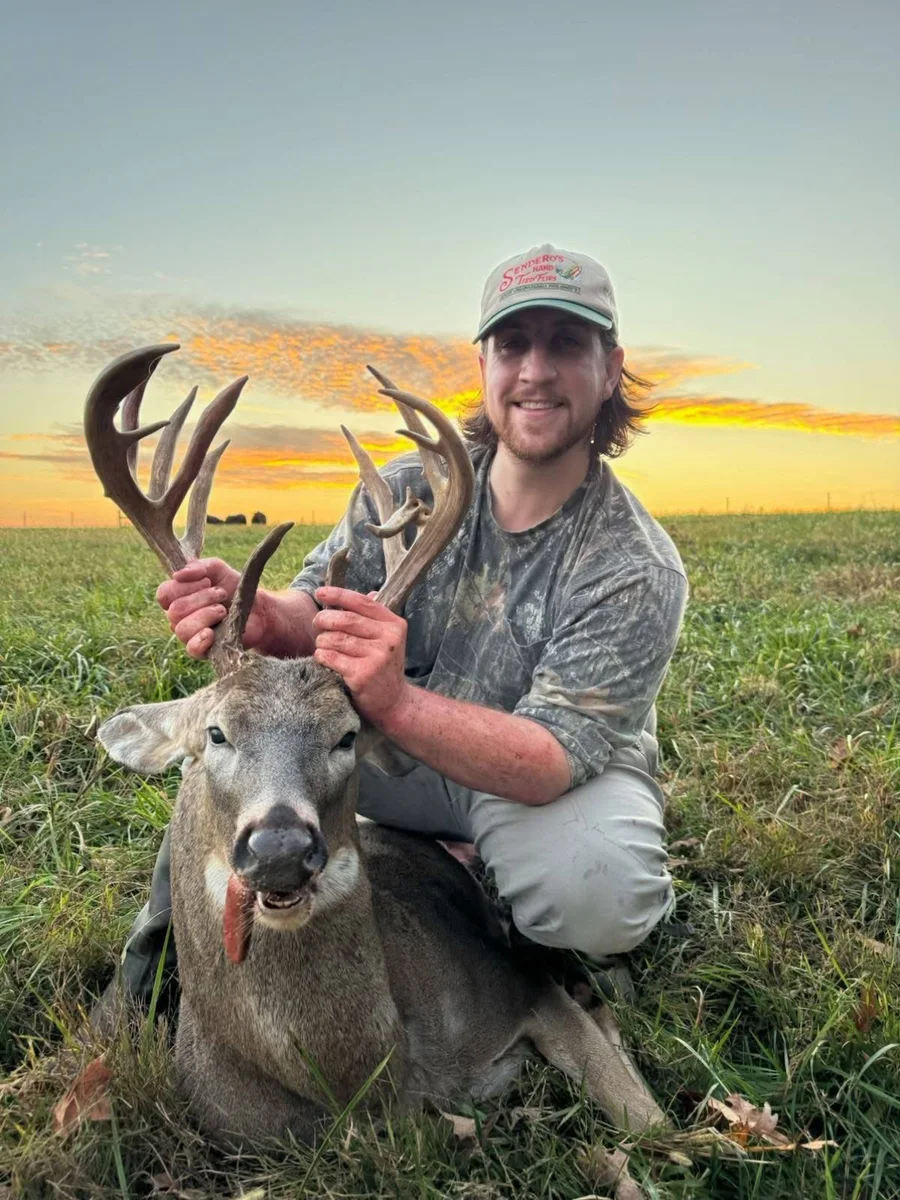 Hunter holding a buck’s antlers in a field at sunset.