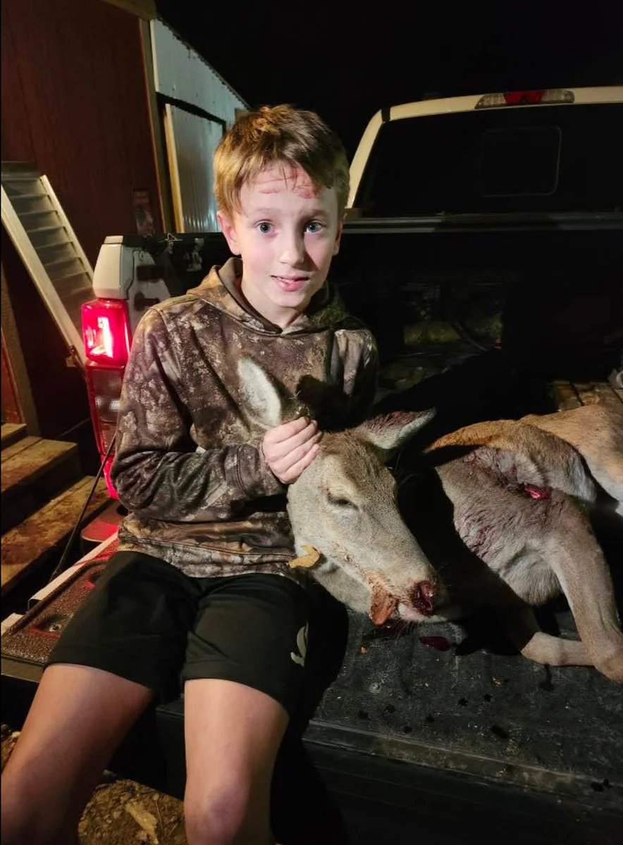 Young hunter sitting on a truck tailgate beside a harvested deer.